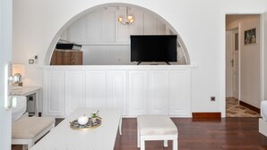 Ett kök med vita köksskåp, en platt-TV, ett matbord med en bänk och en kristallkrona.