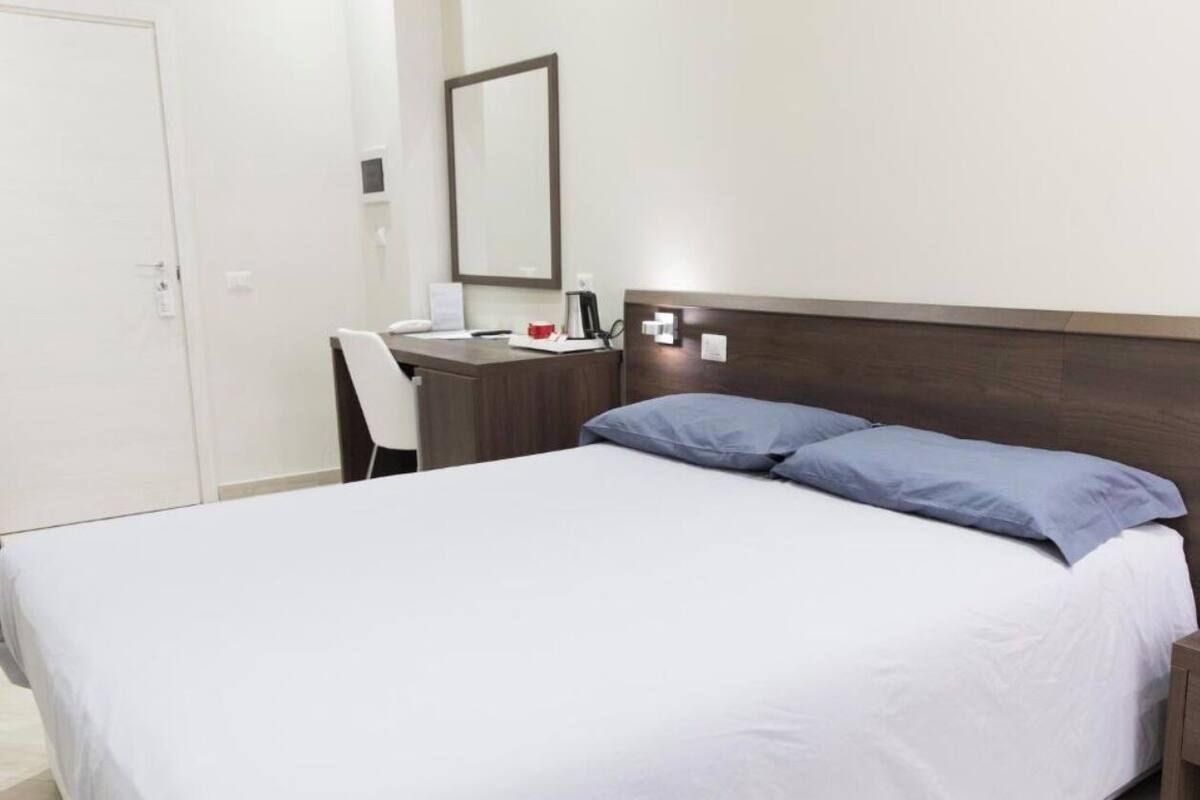 A hotel room with a bed, a desk, a chair, a bedside table, and a mirror.