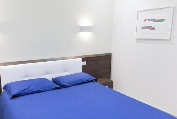 A bedroom with a bed, two pillows, a nightstand, and a wall-mounted light fixture.