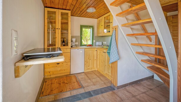 A compact kitchen with wooden cabinets, a built-in dishwasher, and a wooden staircase.