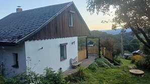 A white house with a sloped roof, a wooden porch, and a view of a lush garden and distant hills.