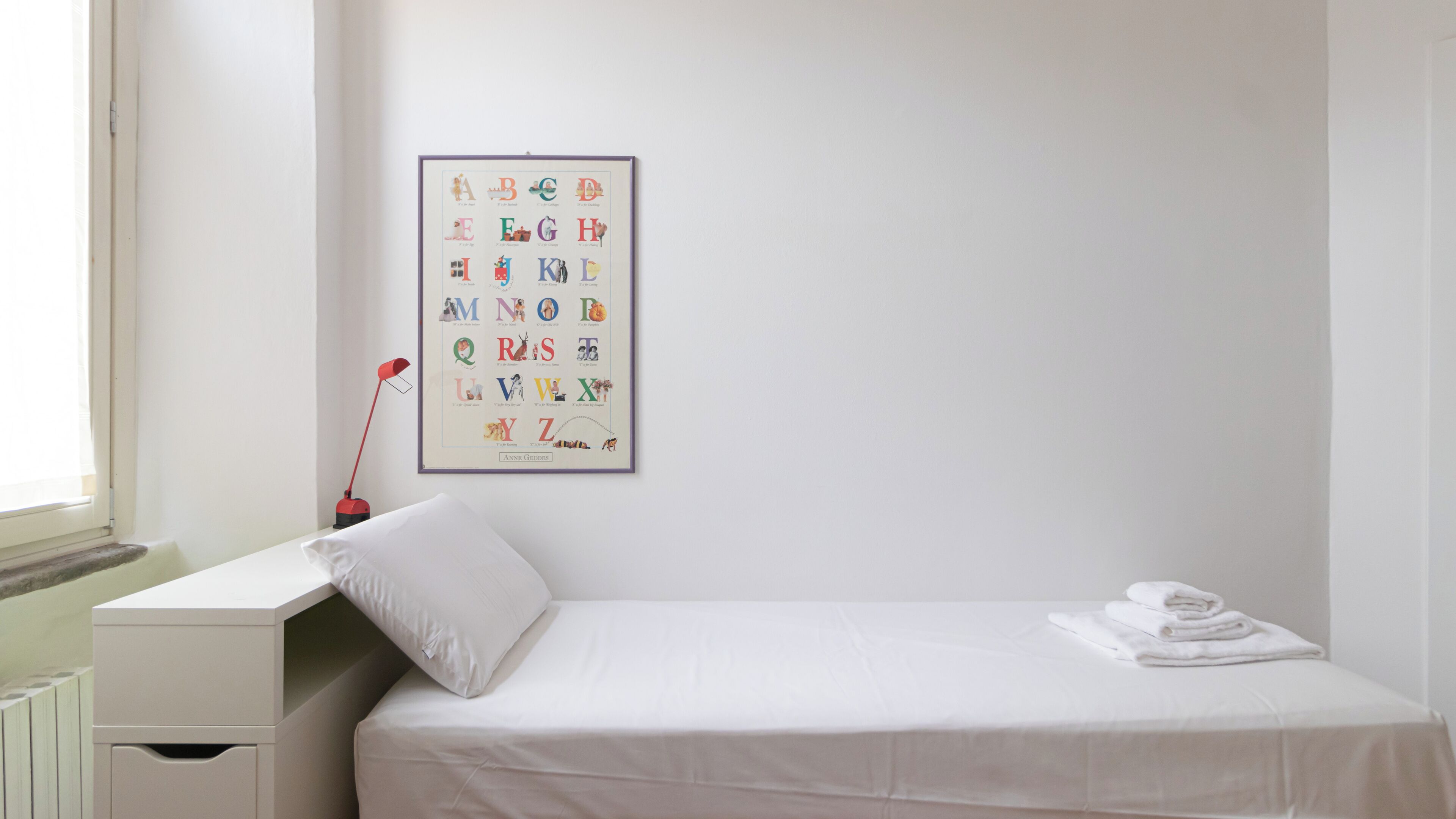 A neatly made bed with a white mattress and pillow, a small bedside table with a lamp, and a framed alphabet poster on the wall.