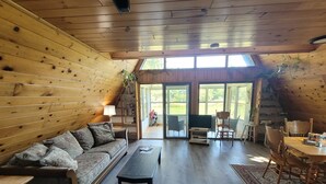 A cozy, wooden-floored living room with a sofa, chairs, a coffee table, and a TV. Large windows provide natural light and a view of the outdoors. - A Frame cabin with a short stroll to a gorgeous sandy beach! (Beaver Island)