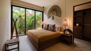 A bedroom with a large bed, a wooden chair, a bedside table with a lamp, a fan, and a view of greenery through a sliding door.