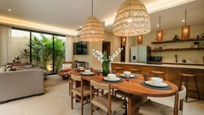 A modern living room with a sofa, dining table, and chairs, a kitchen area with a refrigerator, microwave, and shelves, and large windows with plants.
