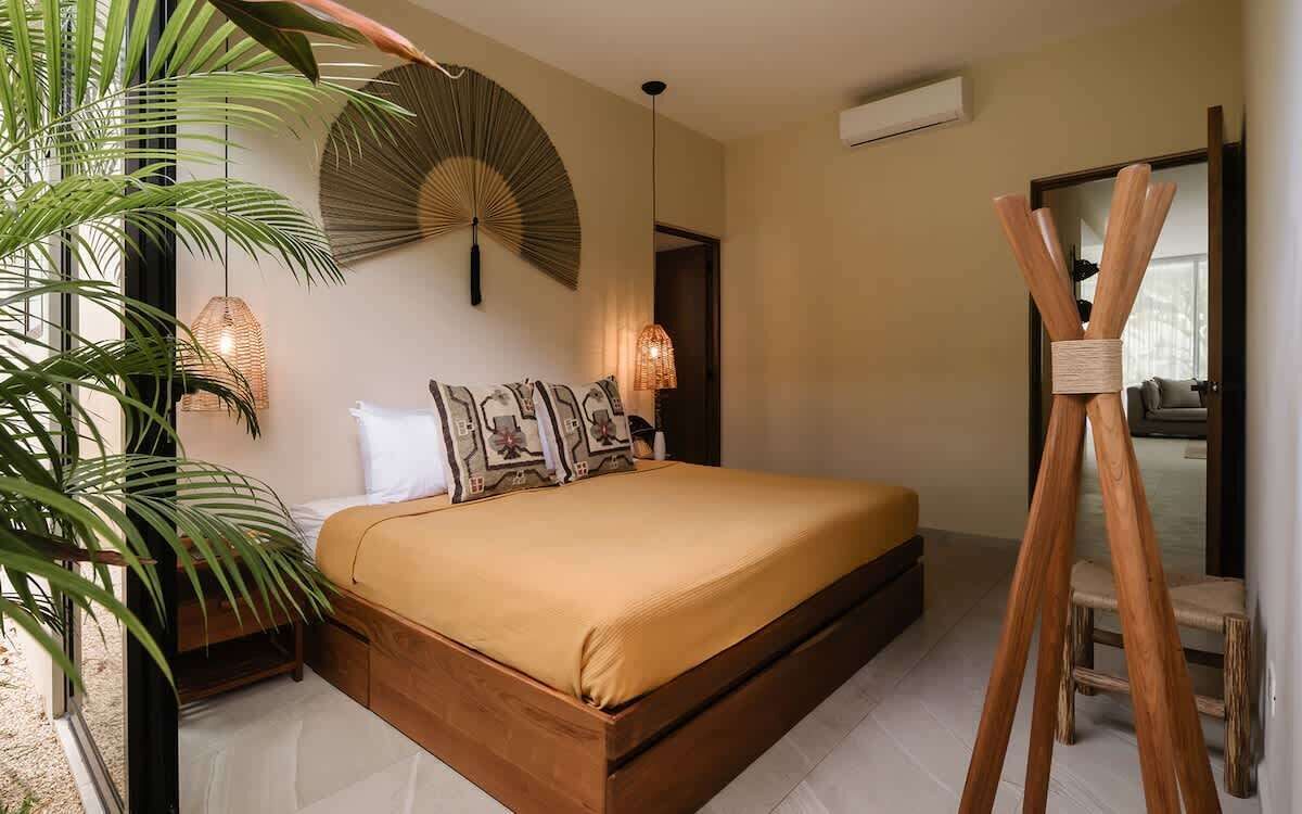 A bedroom with a bed, a standing fan, a wooden chair, and a large plant.