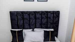 A neatly made bed with a white mattress and a dark headboard. There are two pillows, one black with a yellow stripe and one white. Two framed artworks are hung on the wall above the bed.