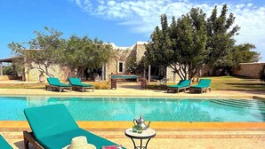 Un espace aménagé au bord de la piscine, avec des chaises longues, une petite table sur laquelle sont posés un chapeau et un magazine, et un bâtiment en pierre en arrière-plan.