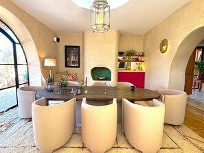 Un coin repas comprenant une table ronde, six chaises, un lustre, une cheminée et un espace cuisine avec des meubles rouges et un plan de travail vert.