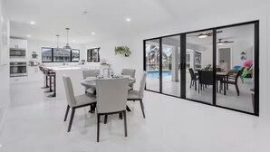 Eine moderne Küche mit Kochinsel, einem Essbereich mit rundem Tisch und Blick auf einen Swimmingpool durch große Glasüren.