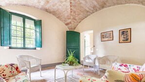 Ein Zimmer mit hoher, gewölbter Decke, einem Fenster mit grünen Rollläden, Korbsesseln, einem floralen Sofa, einem kleinen Tisch mit einer Pflanze und zwei gerahmten Bildern an der Wand.