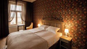 A bedroom with a large bed, two bedside tables with lamps, a chair, a window with curtains, and a floral wallpaper.
