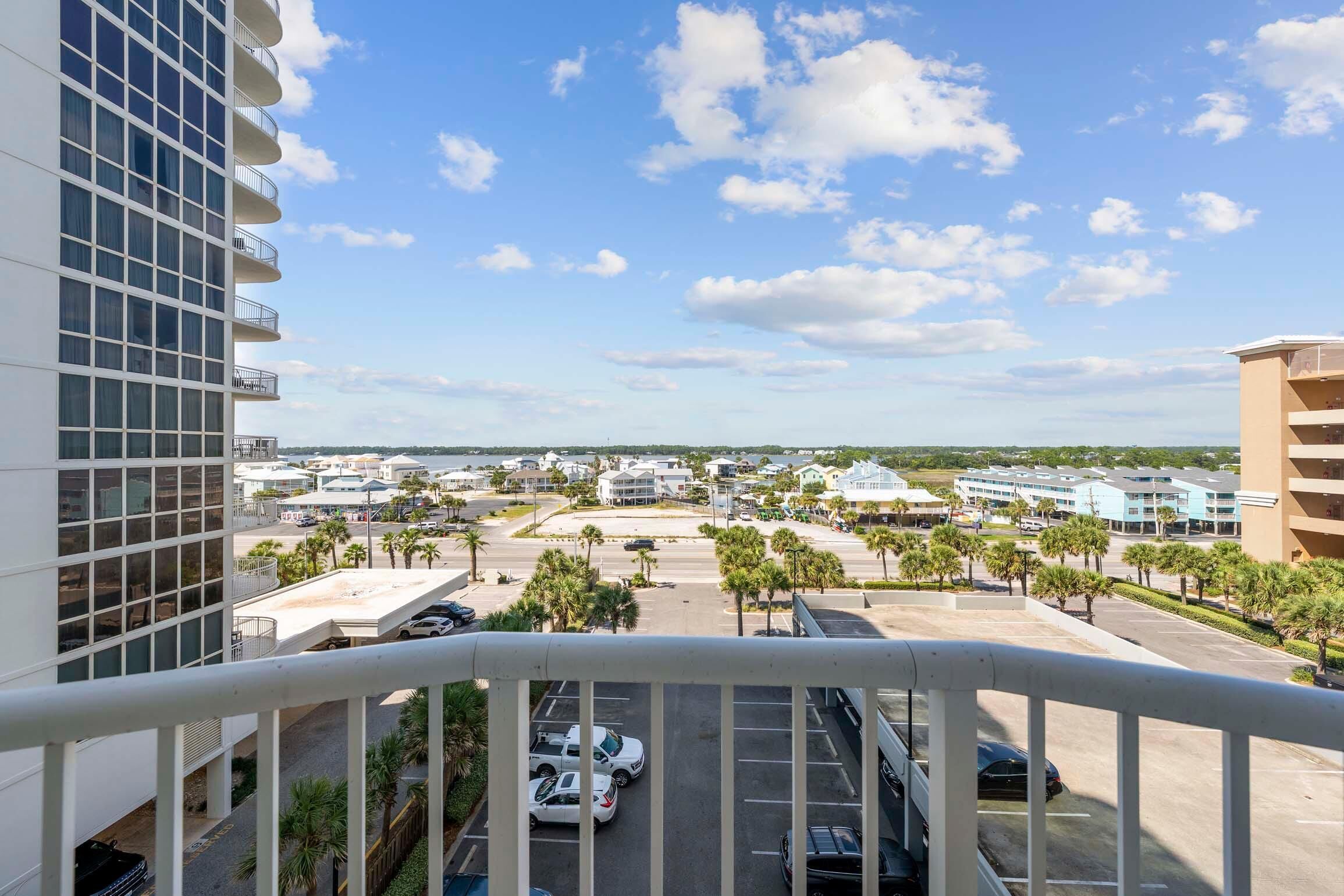 A high-rise hotel with multiple balconies, a parking area, and a view of a coastal town with buildings and palm trees.