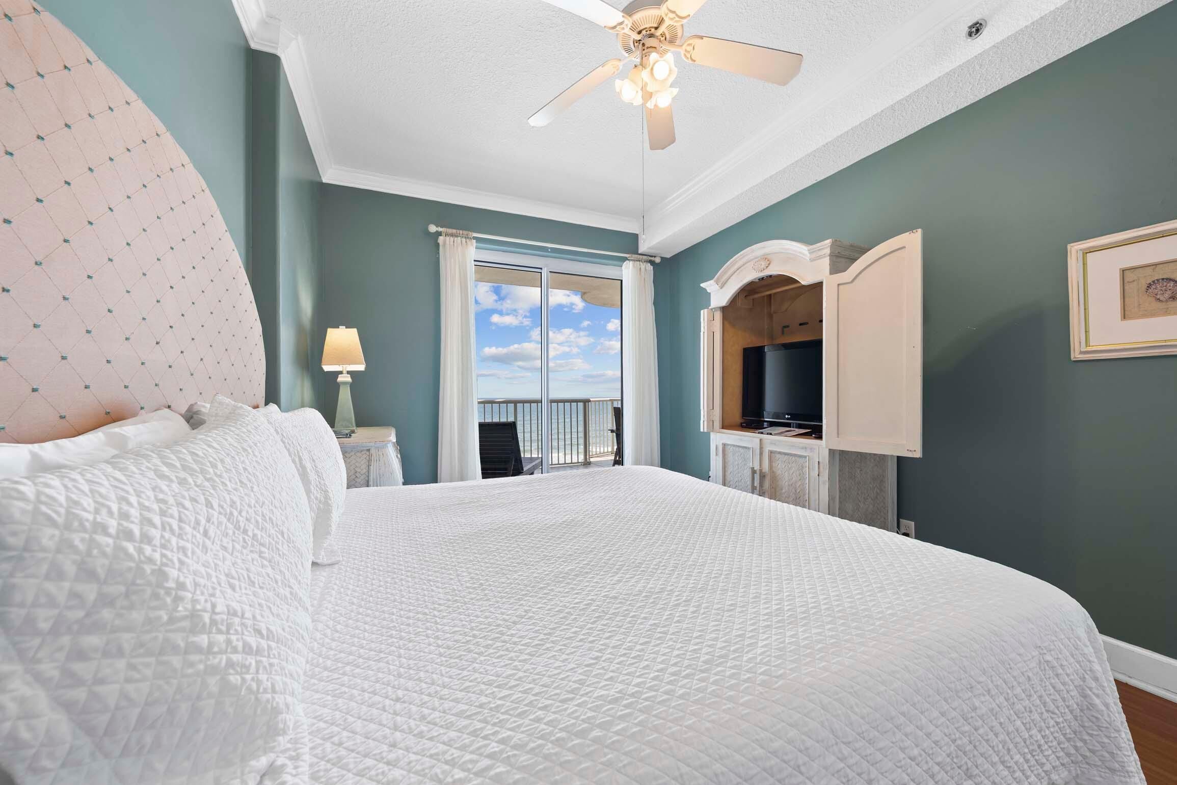 A bedroom with a large bed, a ceiling fan, a TV, and a view of the sky through the window.