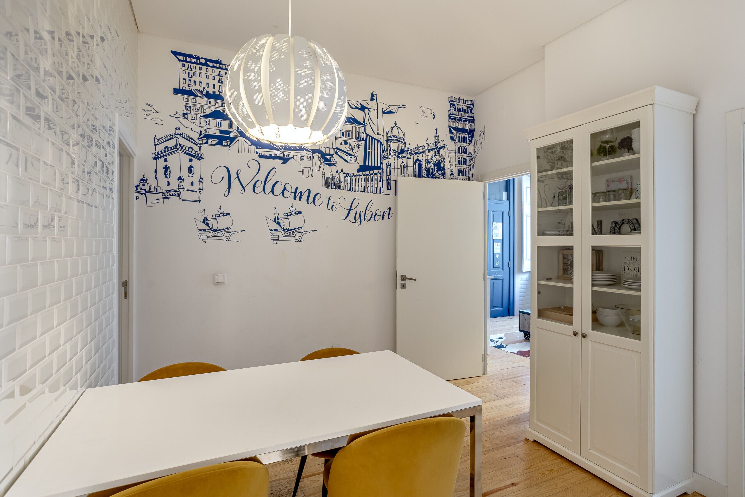 A dining area with a white table, yellow chairs, and a wall with blue illustrations and the text 'Welcome to Lisbon'.