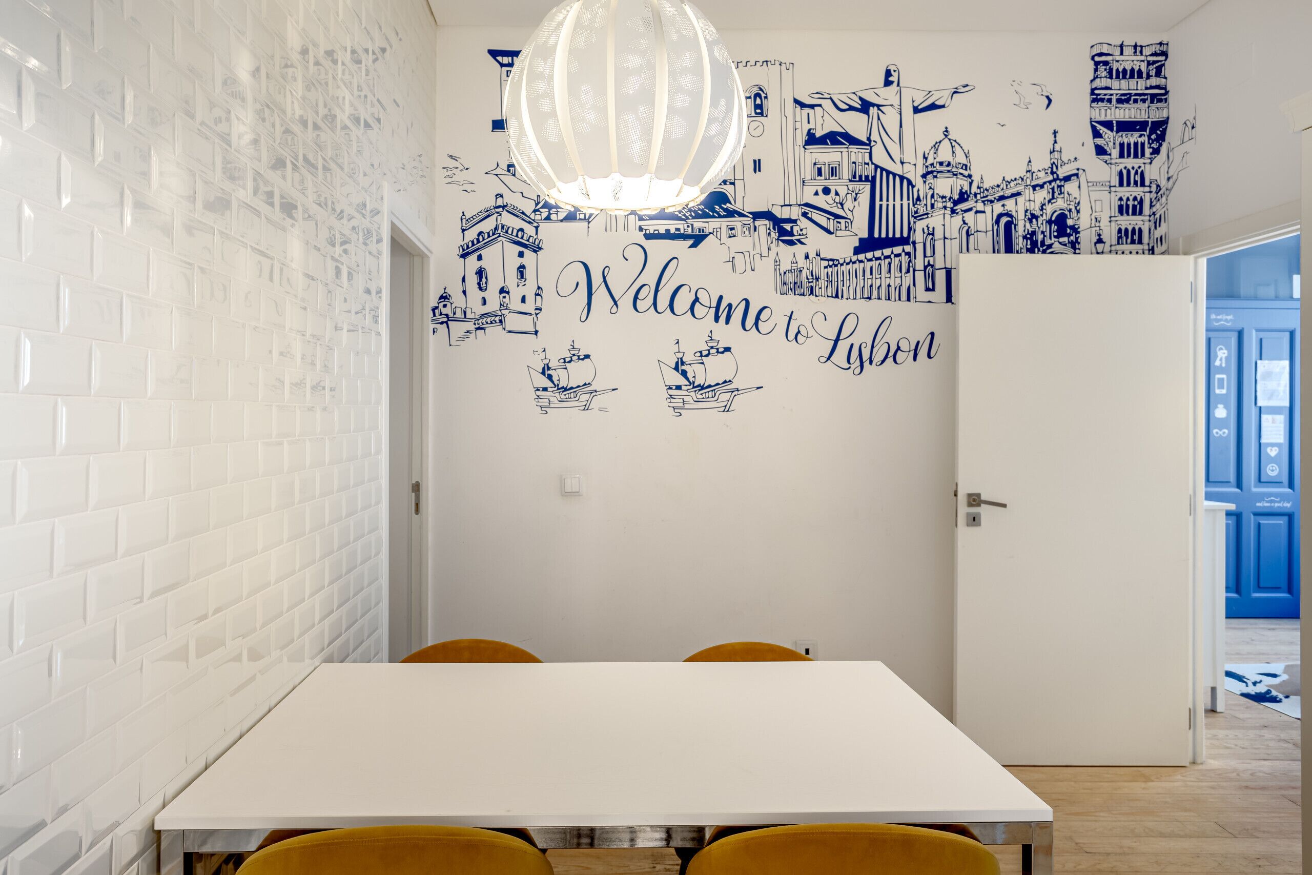 Ein Essbereich mit einem weißen Tisch, gelben Stühlen und einer Wand mit einem Stadtbild-Mural und dem Schriftzug 'Welcome to Lisbon'.