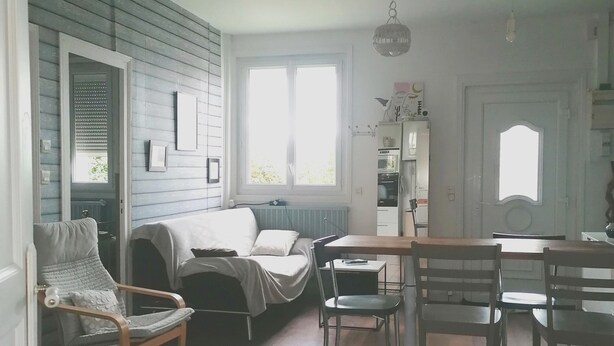 A room with a wooden floor, a sofa, a dining table with chairs, a hanging light fixture, and a window.
