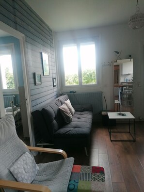 A living room with a sofa, armchair, and a small table. There is a window with a view of greenery outside. - Holiday Home 'Maison' with Private Terrace, Private Garden and Wi-Fi (Royan)