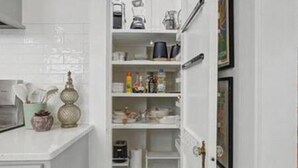 Una cocina con armarios blancos, una encimera con un hervidor, una cafetera y un estante con diversos objetos.