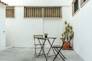 Une petite terrasse extérieure avec une table en bois et deux chaises, un pot de fleurs et des fenêtres grillagées.