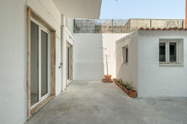 Il s’agit d’un espace extérieur étroit et clos, doté d’un sol en béton, d’une porte et d’une petite fenêtre. Des plantes en pot sont disposées le long du côté droit.