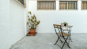 Un pequeño espacio al aire libre con una mesa y sillas, una planta en maceta y ventanas con barrotes en una pared blanca.