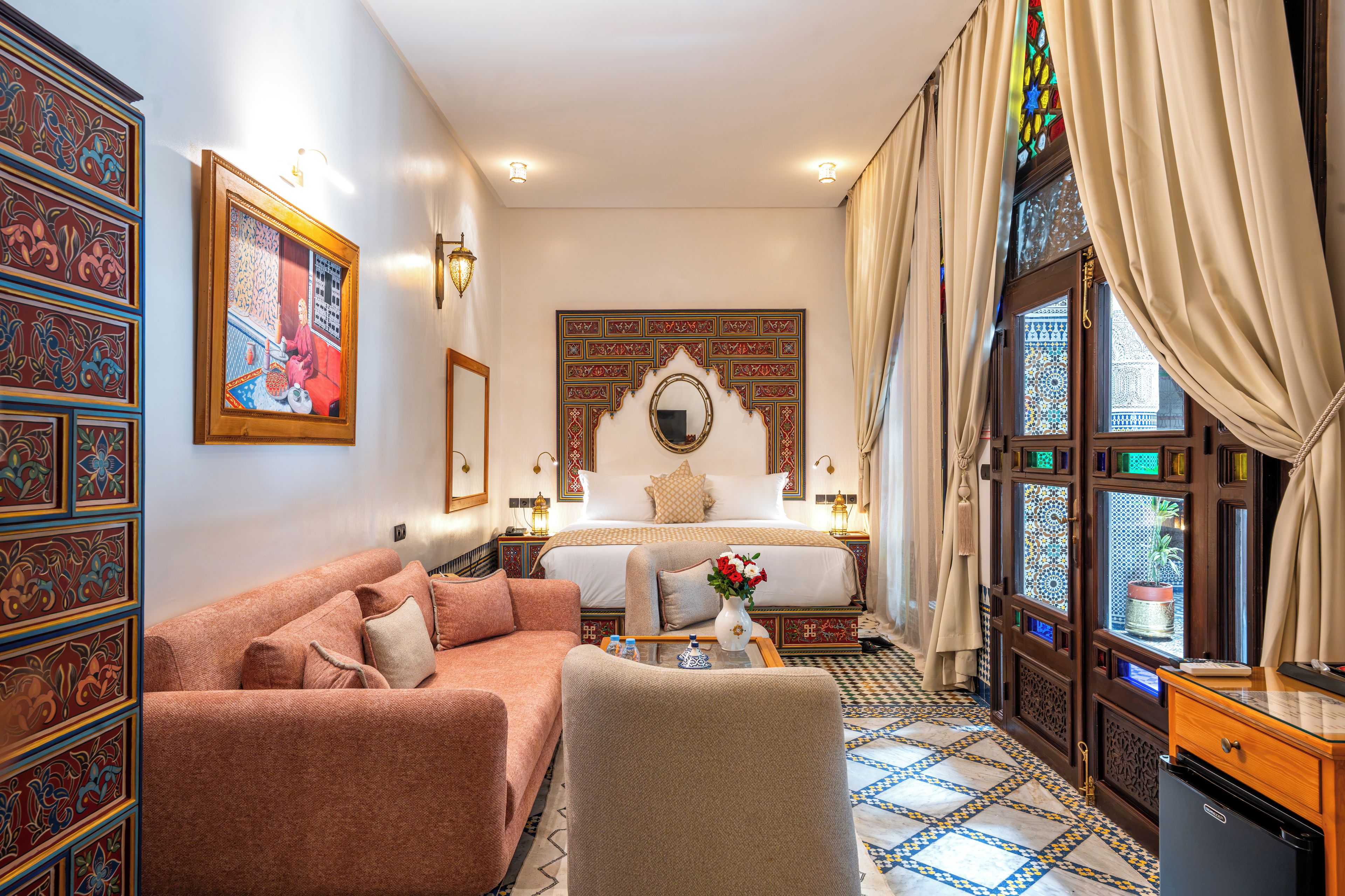 Quarto tradicional com cama, sofá, espelho decorativo e vista pela janela com cortinas.