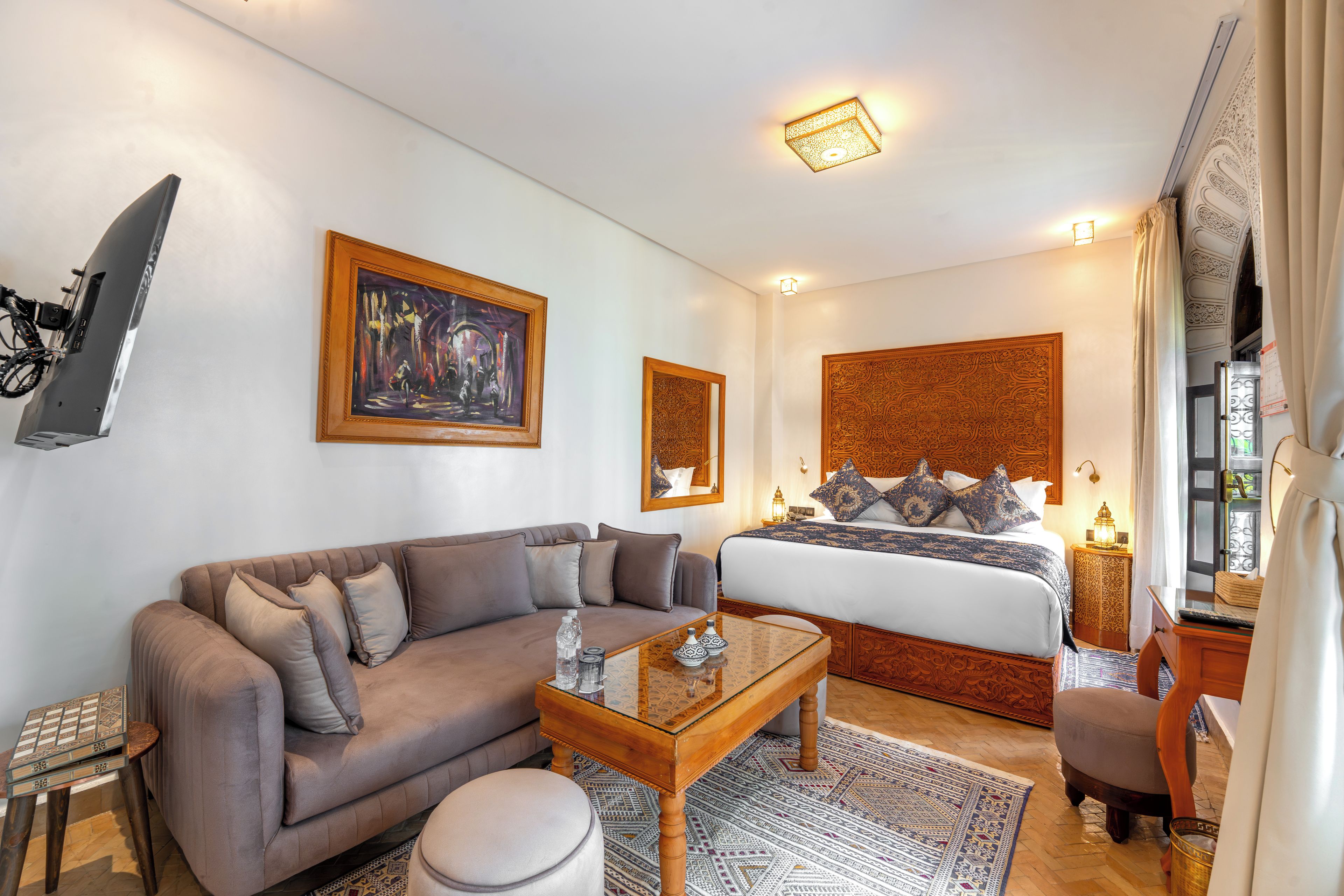 Een moderne hotelkamer met een bank, een bed met decoratieve hoofdbord, een flatscreen televisie aan de muur en een klein tafeltje met glazen blad.