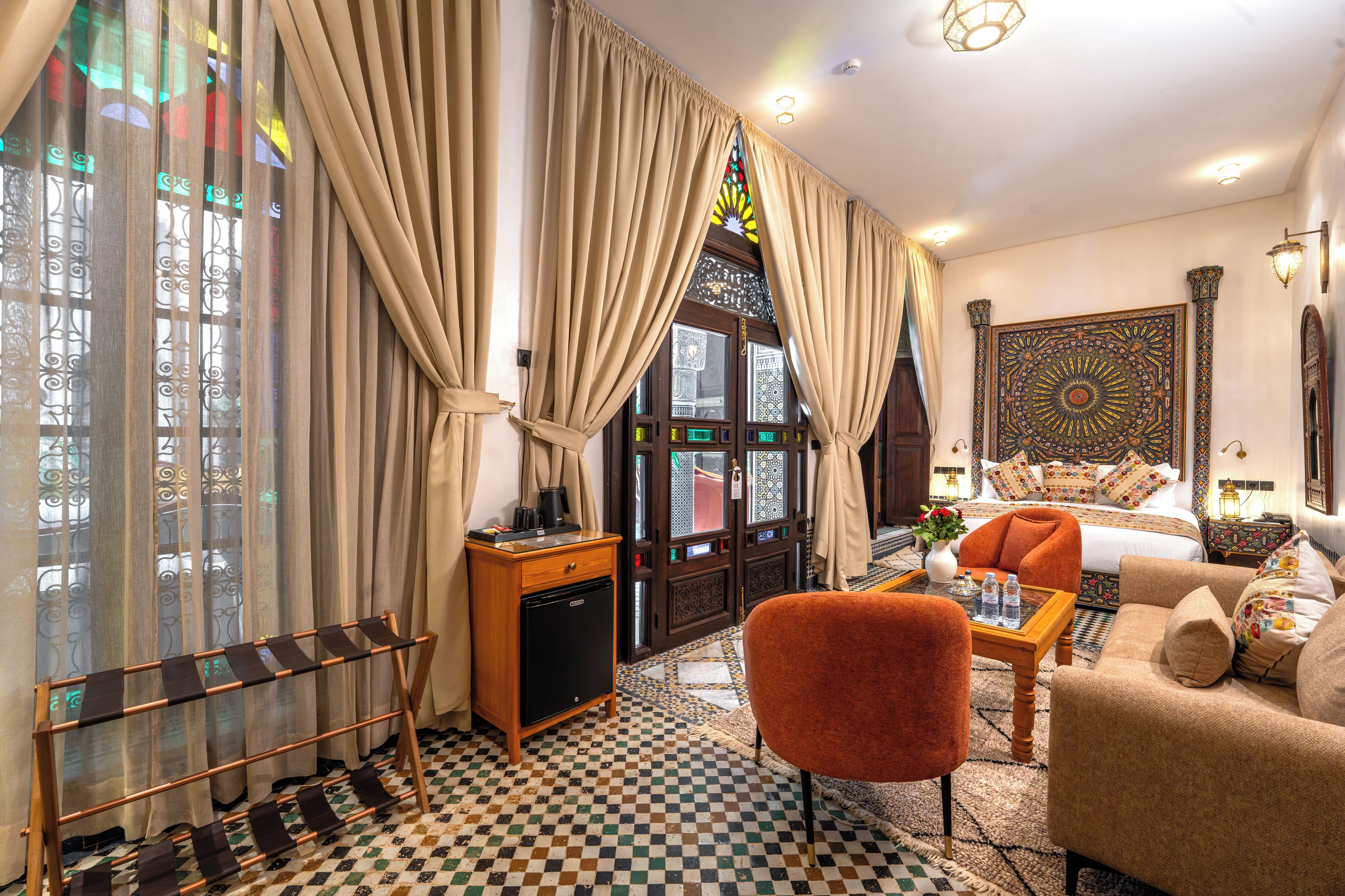 Een hotelkamer met een zwart-wit schakeerdvloer, een bank, een salontafel, een kleine koelkast en gordijnen.