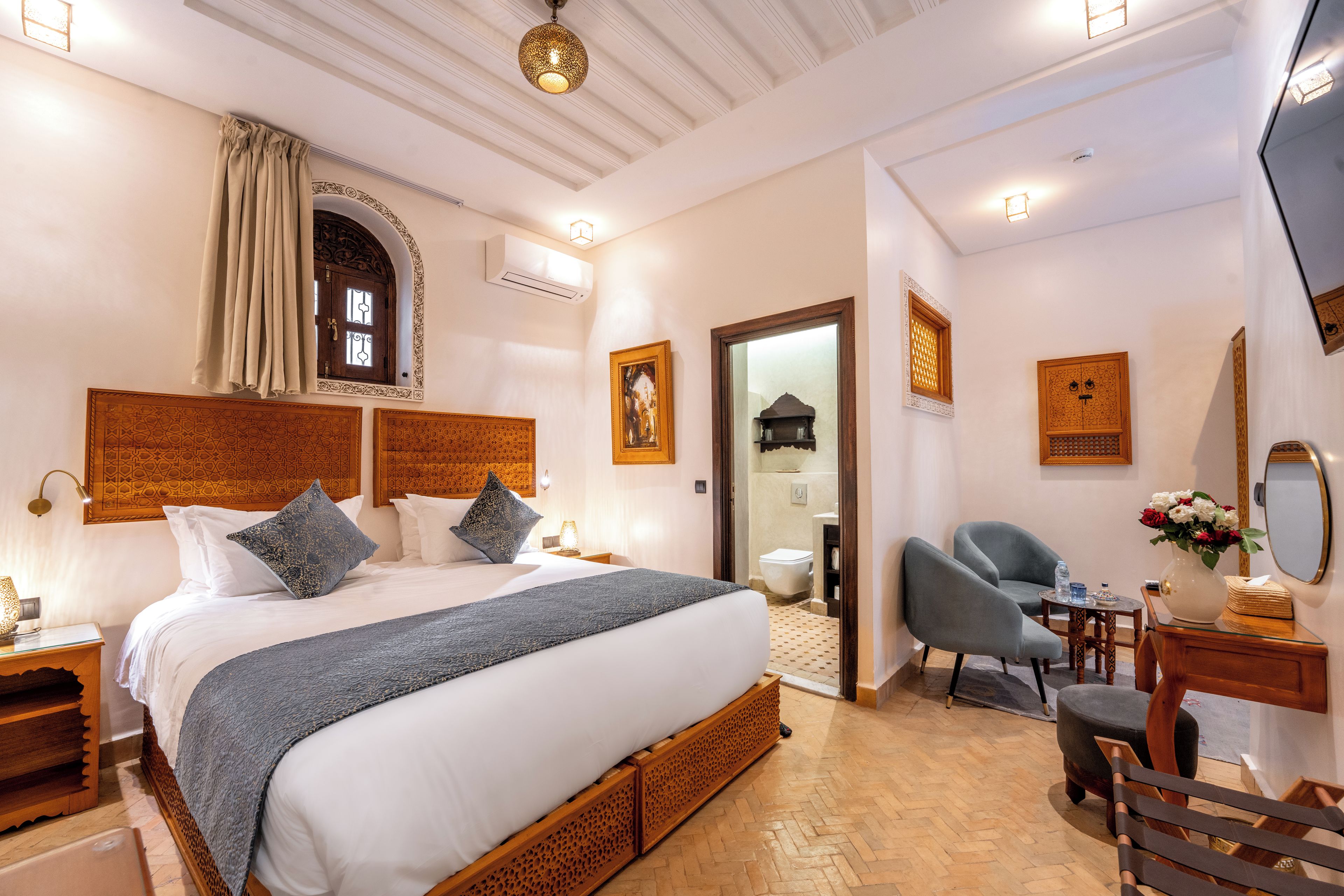 A well-lit bedroom with a large bed, a sitting area with a chair, a small table, and a mirror.