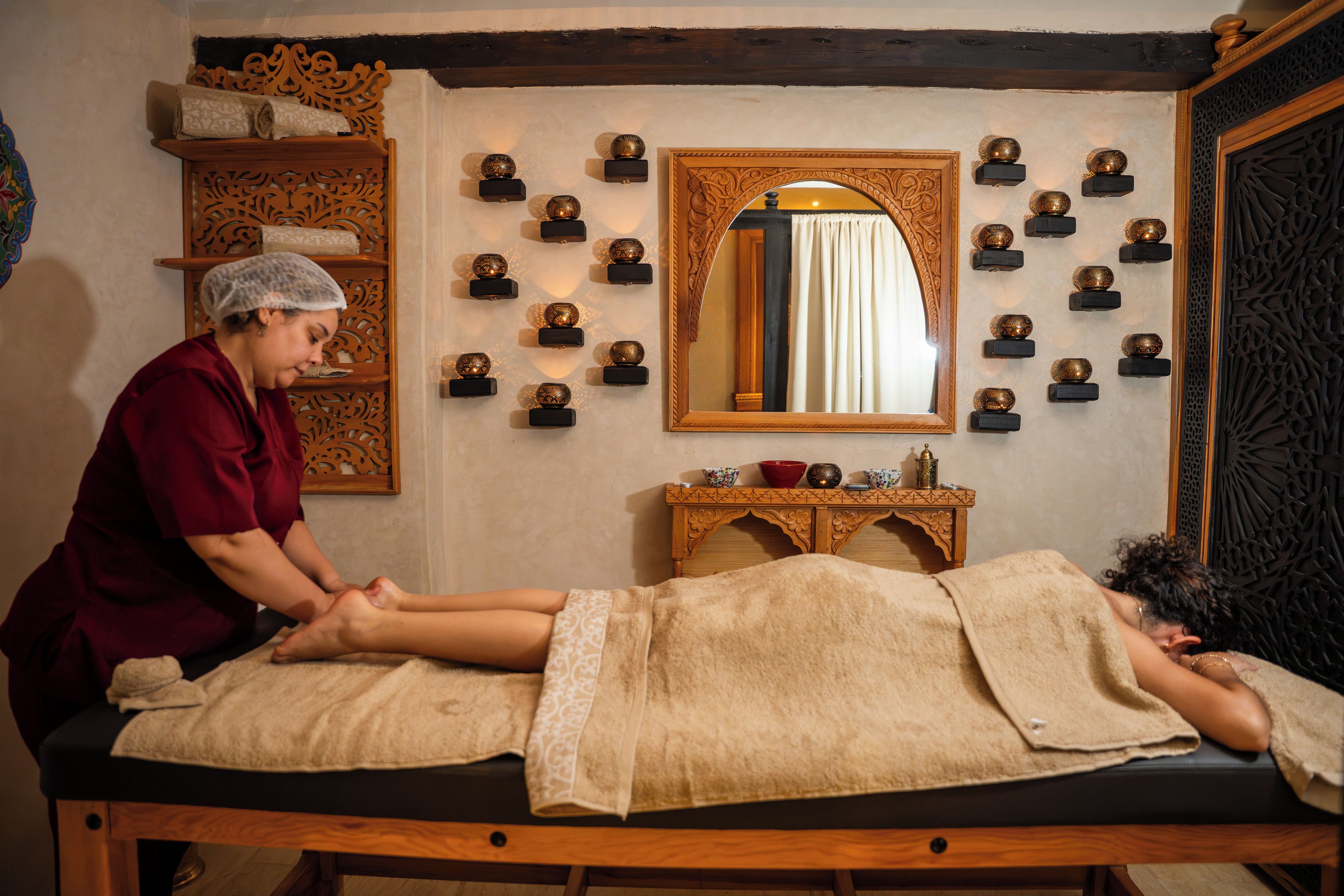 A massage therapy room with a wooden massage table, a wooden cabinet with decorative items, and a mirror on the wall.