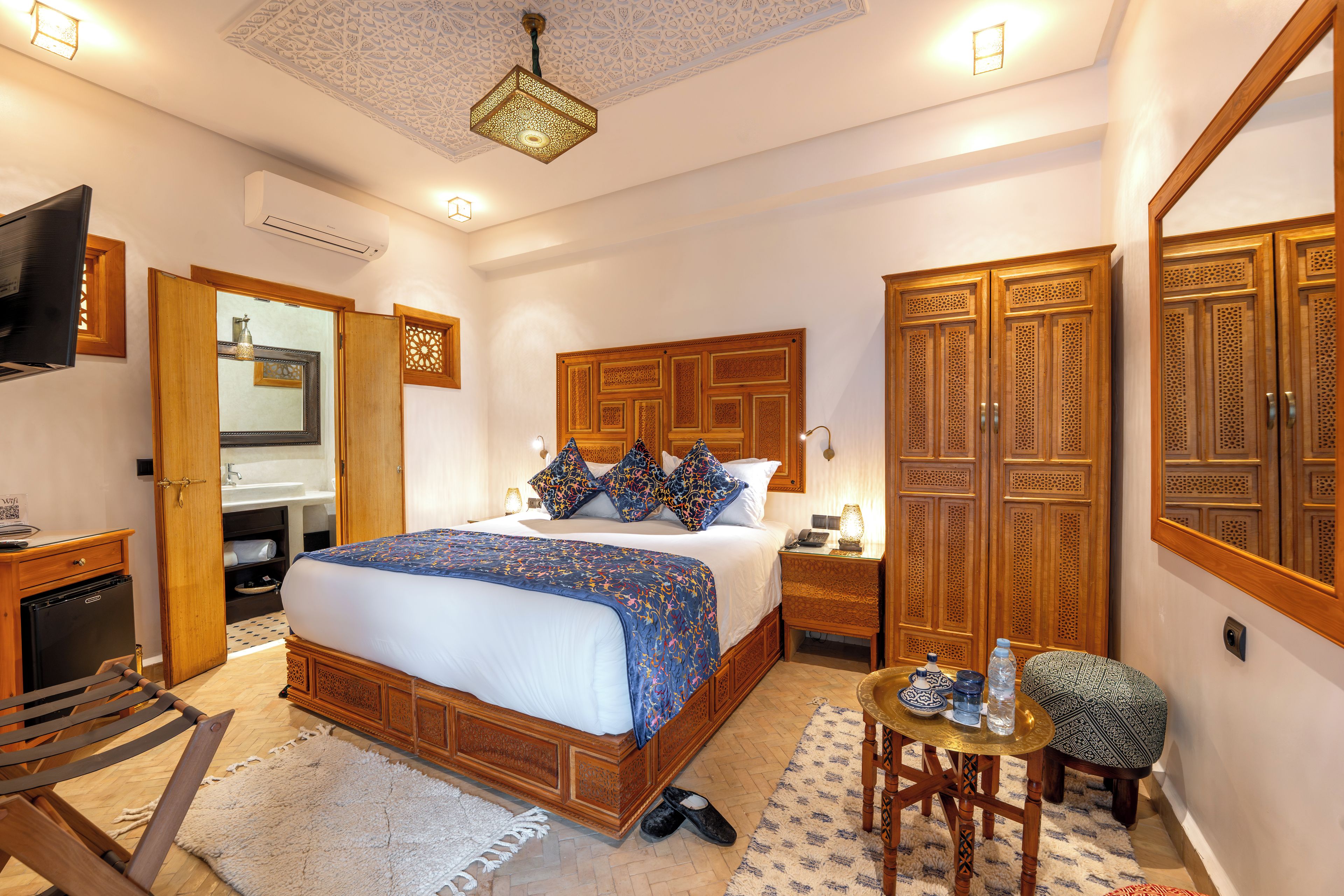 A bedroom with a large bed, a wooden wardrobe, a desk with a chair, and a small table with decorative items.
