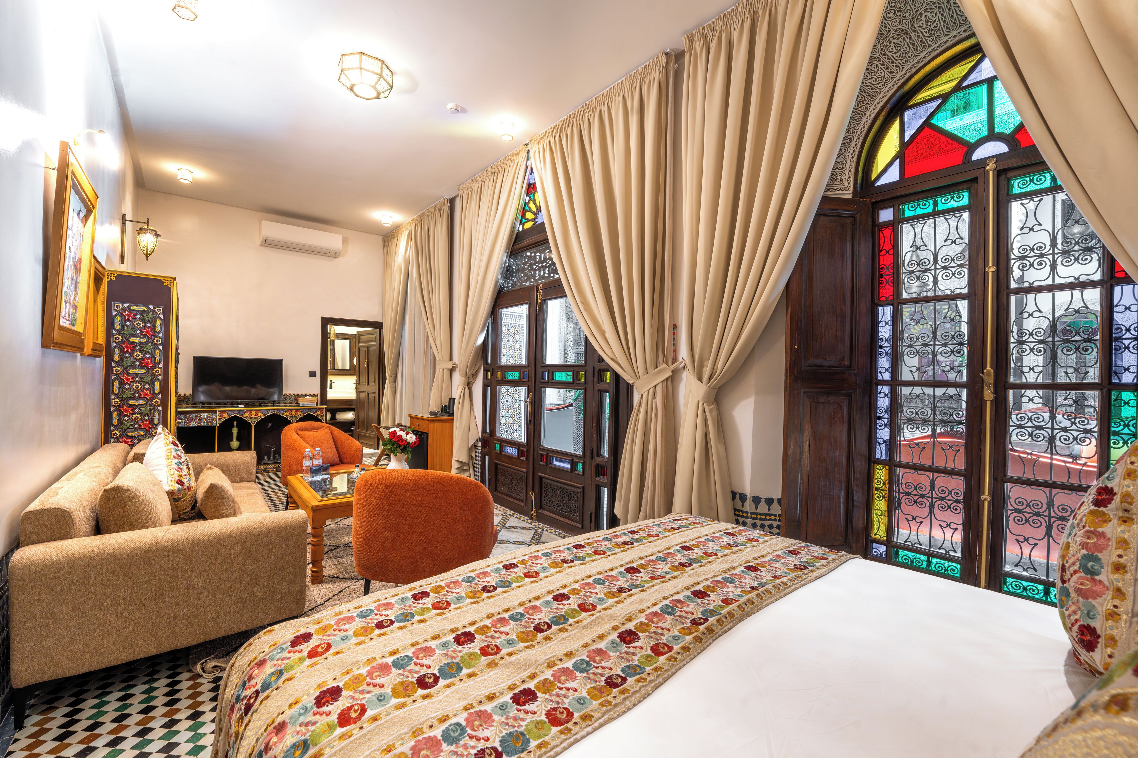 A hotel room with a large bed, a sofa, a TV, and a view of a colorful building through a stained glass window.