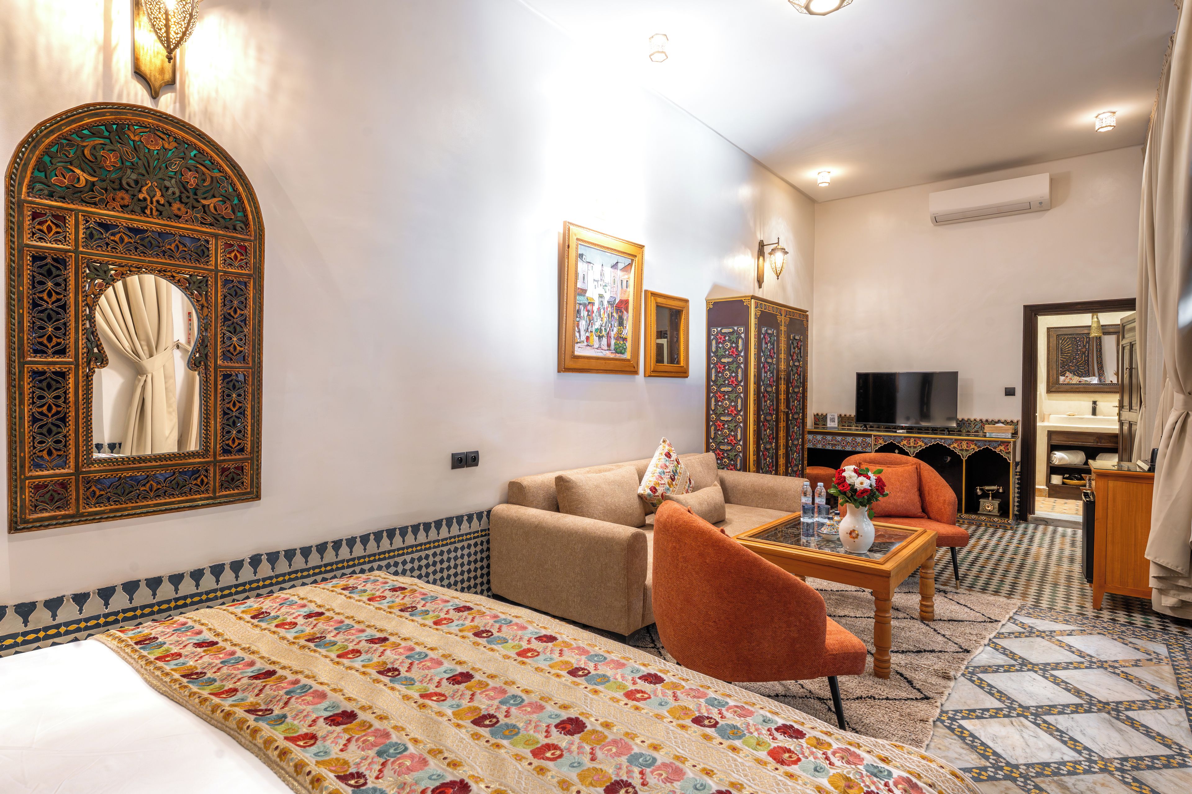 Quarto de hotel com cama, sofá, mesa de centro, televisão e banheiro visível através de uma porta aberta.