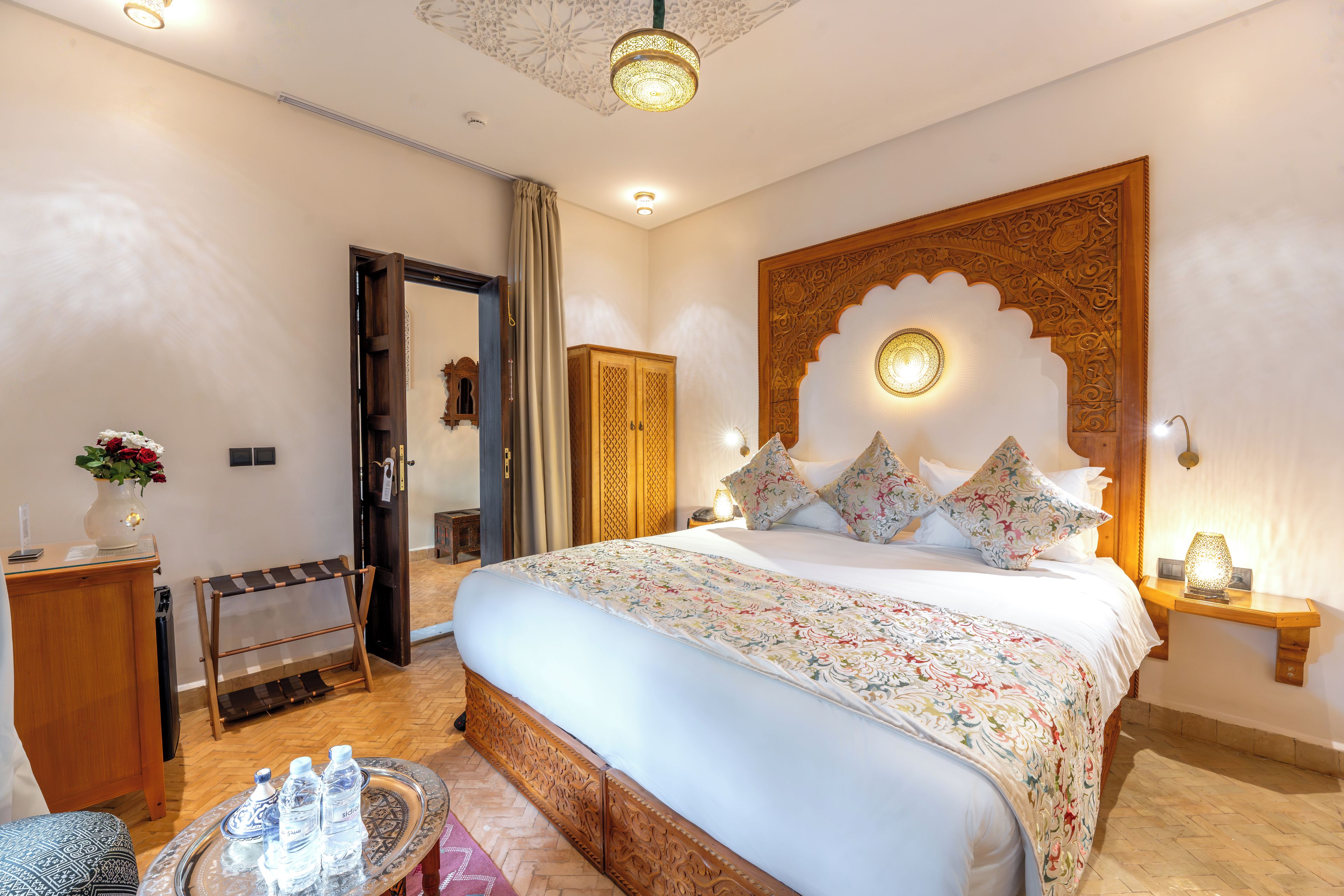Ein stilvoll eingerichtetes Hotelzimmer mit einem großen Bett, einem hölzernen Kopfteil, einem Nachttisch mit Lampe, einem kleinen Tisch mit Wasserflaschen und einer Tür zu einem weiteren Raum.