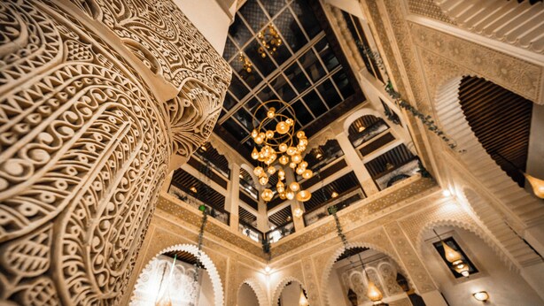 An ornate interior with intricate architectural details, including carved columns and arches, and a central chandelier. - Riad Joy (Fes)