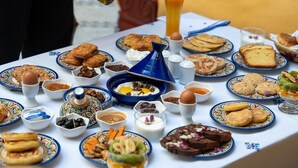 A table set with various breakfast items, including pastries, fruits, and beverages, in a room with a sofa and a patterned tablecloth.