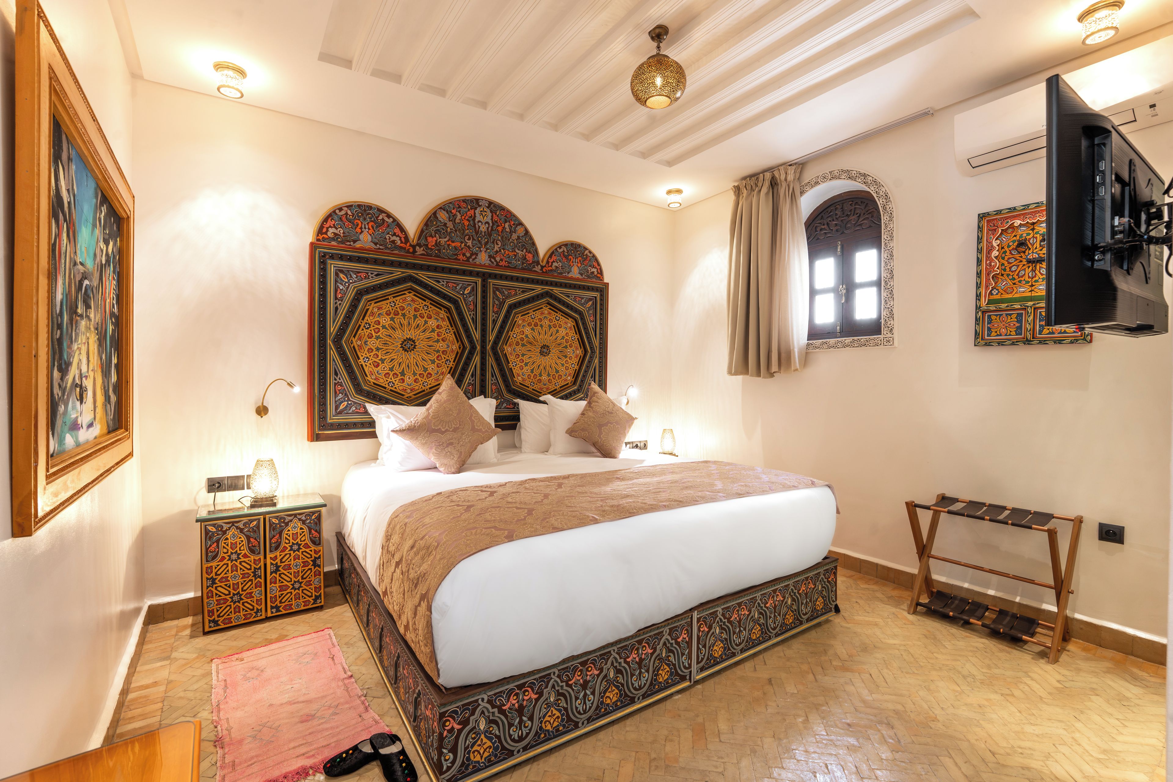 A well-lit bedroom with a large bed, decorative headboard, and a television mounted on the wall.