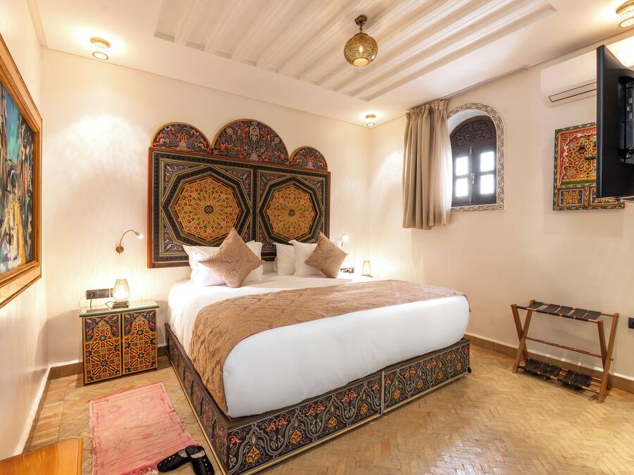 A well-lit bedroom with a large bed, decorative headboard, and a television mounted on the wall.