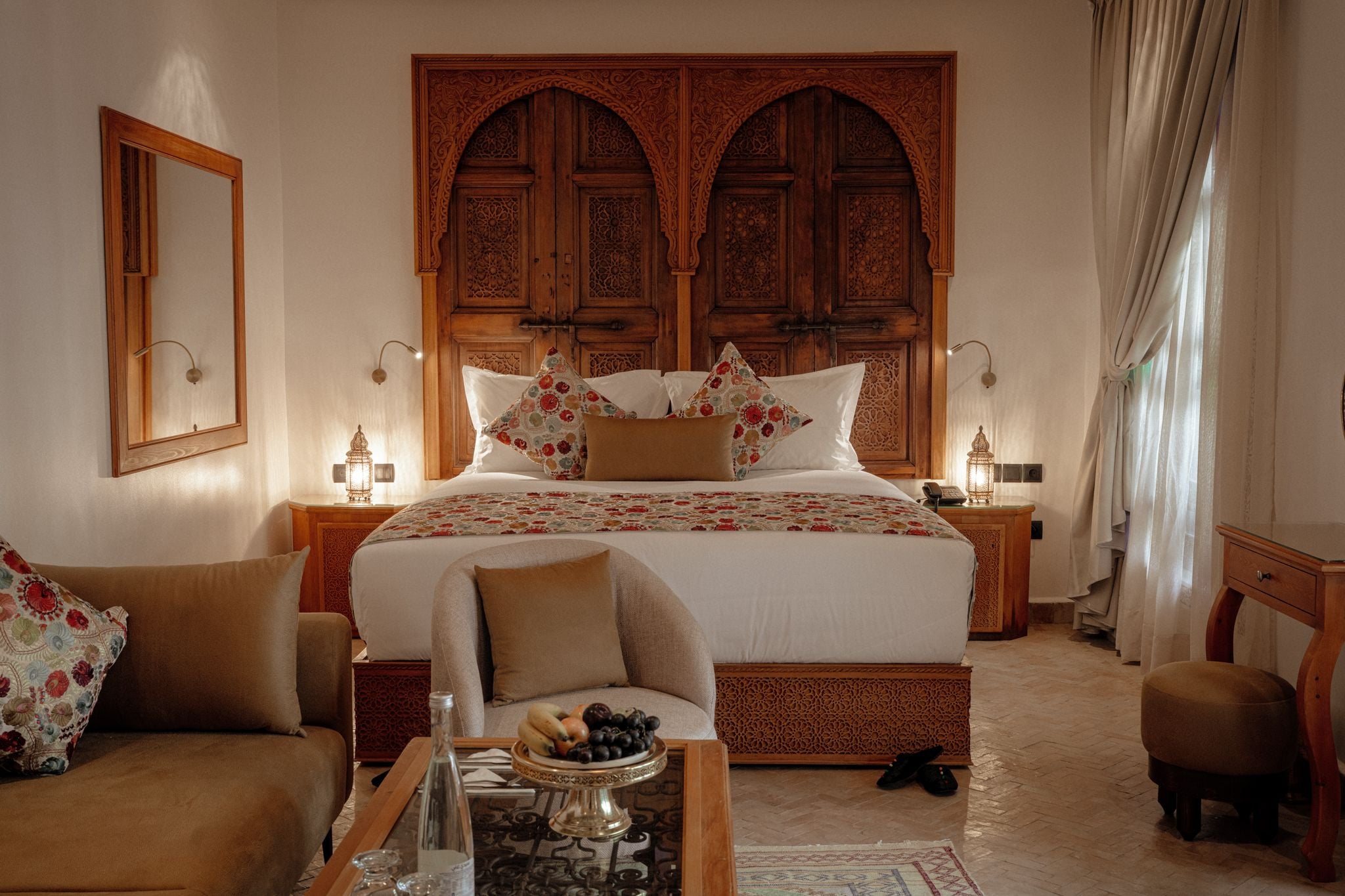A bedroom with a large bed, two nightstands, a chair, a small table with a fruit bowl, and a wooden wardrobe with intricate carvings.