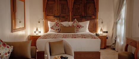 A bedroom with a large bed, two nightstands, a chair, a small table with a fruit bowl, and a wooden wardrobe with intricate carvings.