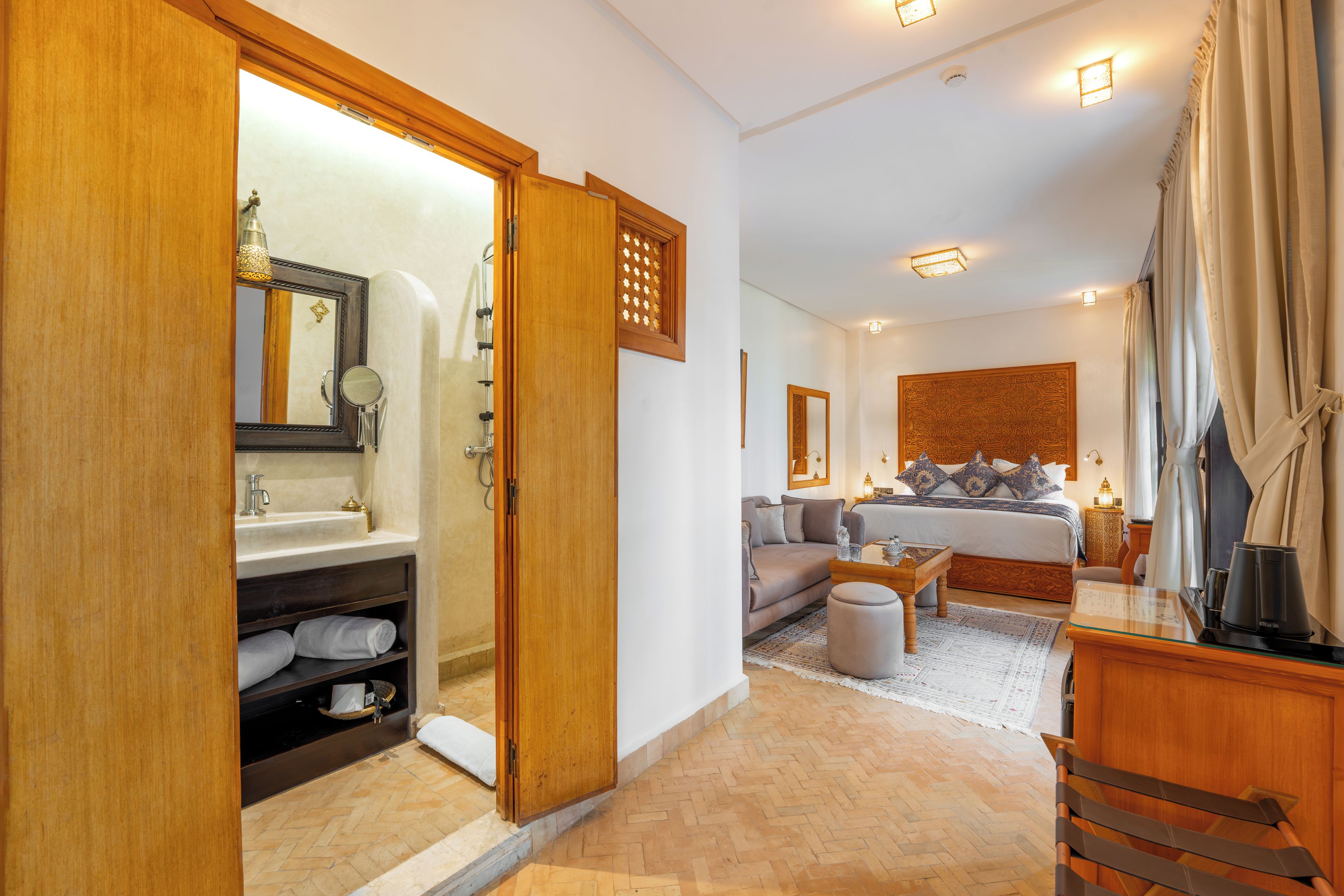 Habitación de hotel con baño, zona de estar con sofá y mesa de centro, y una cama grande con almohadones decorativos.