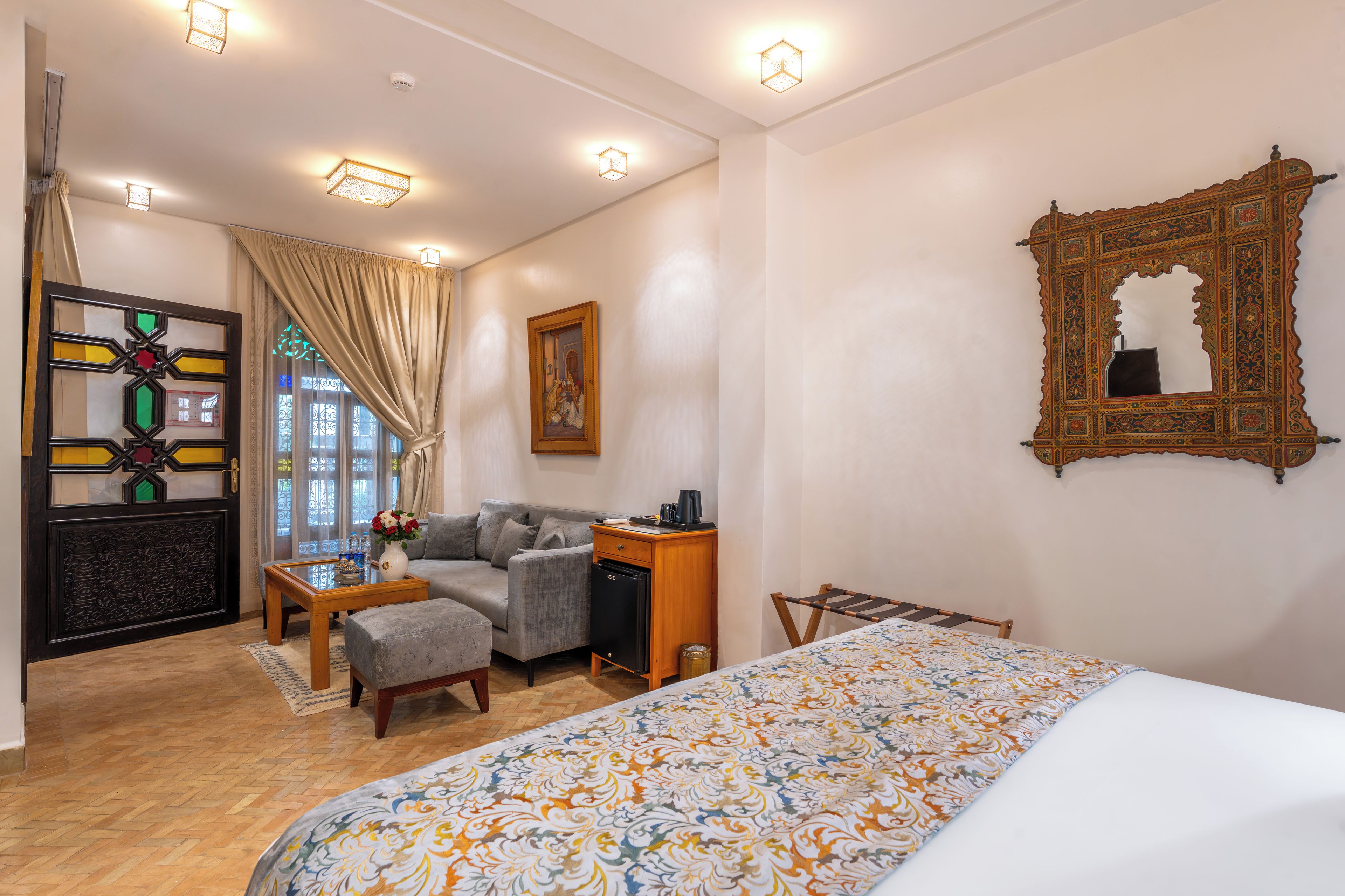 Habitación de hotel con cama, sofá, escritorio, silla, mesita, espejo decorativo y ventana con cortinas.