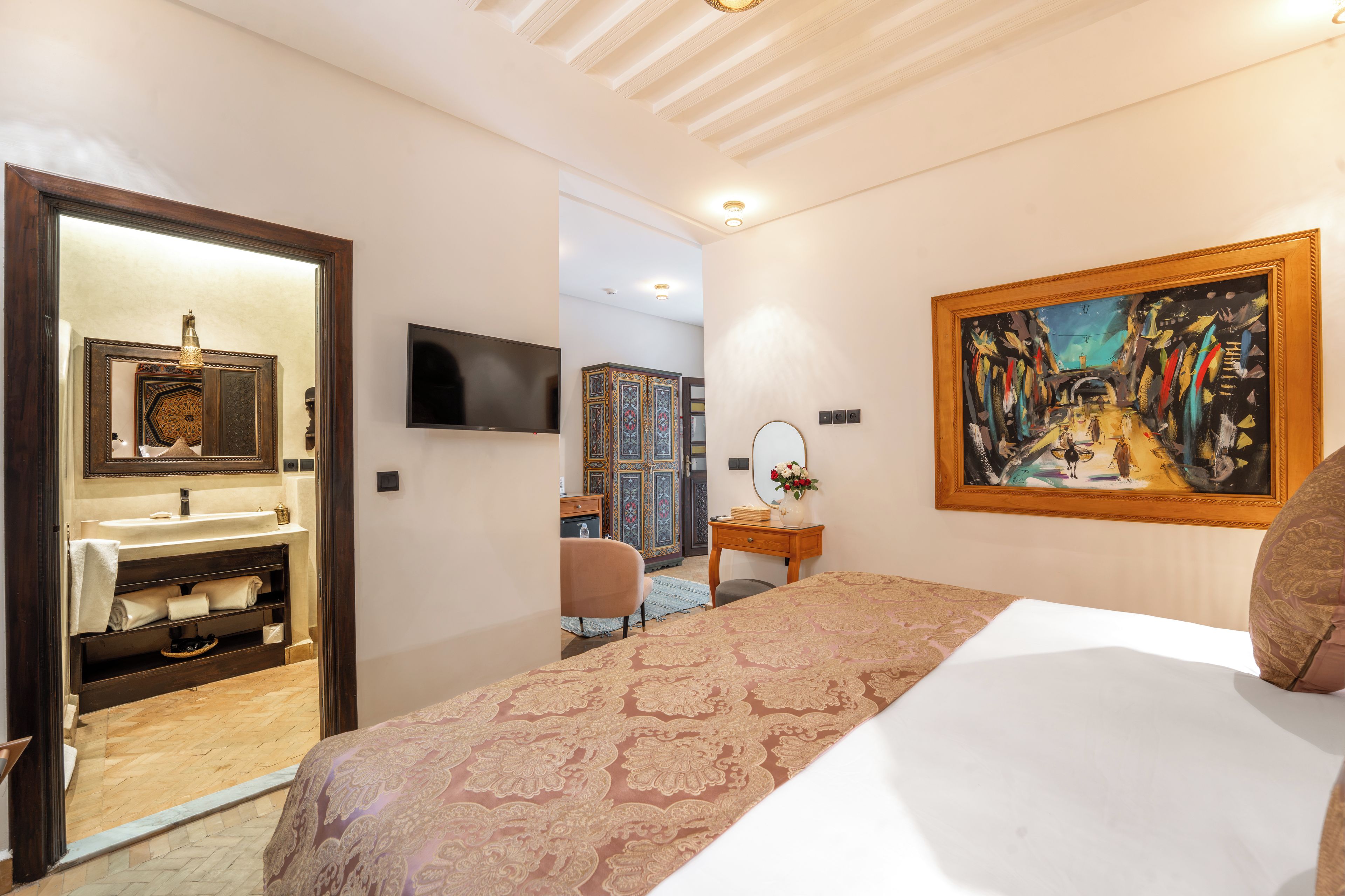 A hotel room with a bed, a television, a painting, and a bathroom with a mirror and sink.