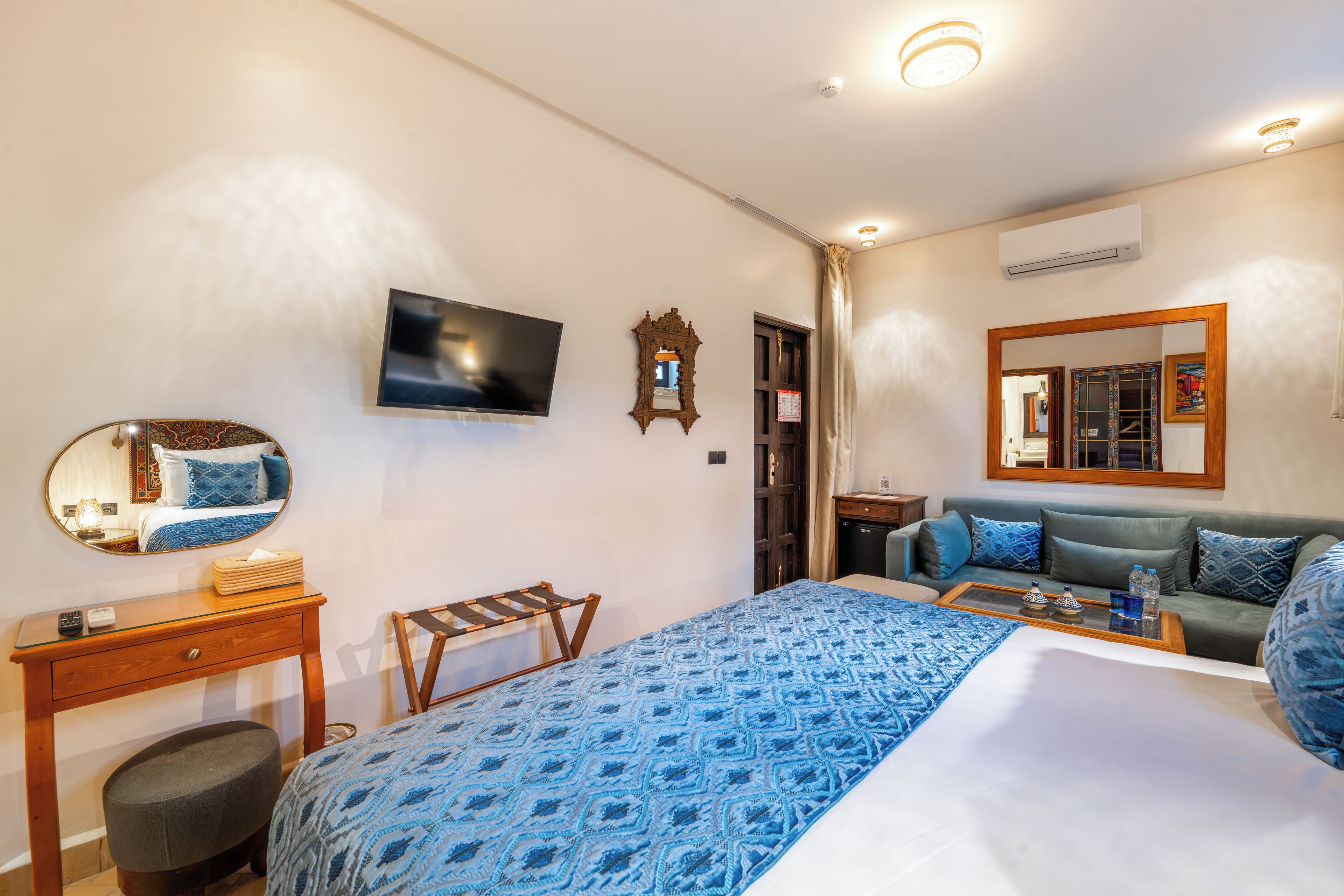 A hotel room with a bed, a television, a mirror, a wooden side table, a sofa, and a small table with a tray.