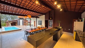 Uma cozinha moderna com uma ilha central, uma área de refeições com cadeiras e uma área de piscina visível por um espaço aberto.