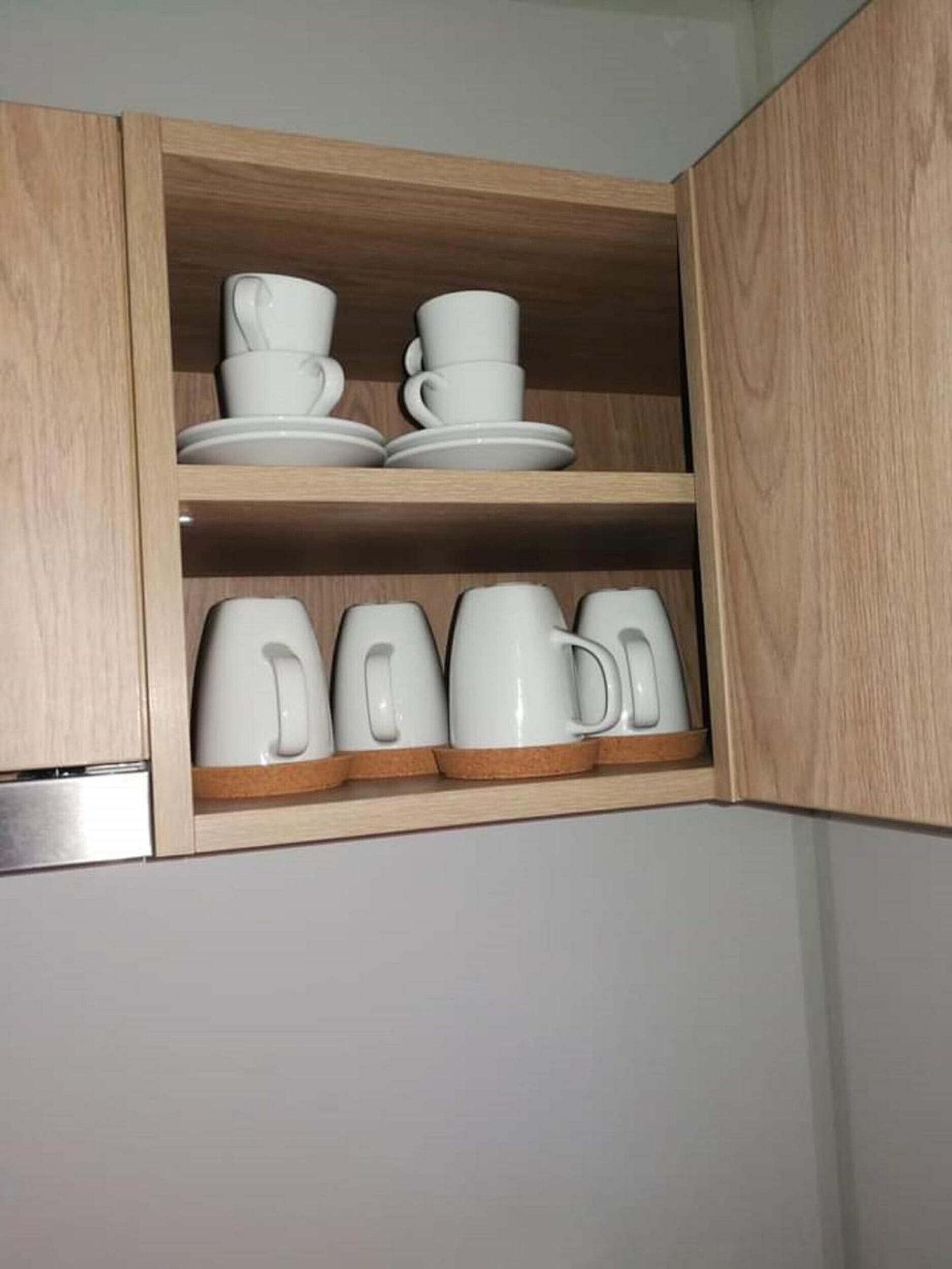 A kitchen cabinet with white ceramic cups and saucers on the upper shelf and white mugs on the lower shelf.