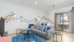 Un salon moderne avec un canapé gris, des fauteuils bleus et roses, une table basse en verre et un tapis bleu. On aperçoit un téléviseur sur la gauche et, au fond, un coin repas avec une table et des chaises.