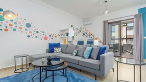 Un salon moderne avec un canapé gris, des coussins bleus et blancs, une table basse en verre et un tapis bleu. Un mur est décoré de motifs circulaires colorés, et une petite étagère présente des objets décoratifs.