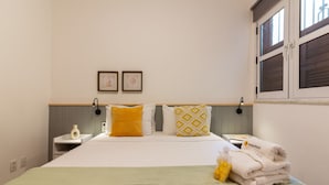 A neatly made bed with a green throw blanket, two pillows with yellow and white patterns, and a stack of white towels labeled "Pineapples" on a bedside table.